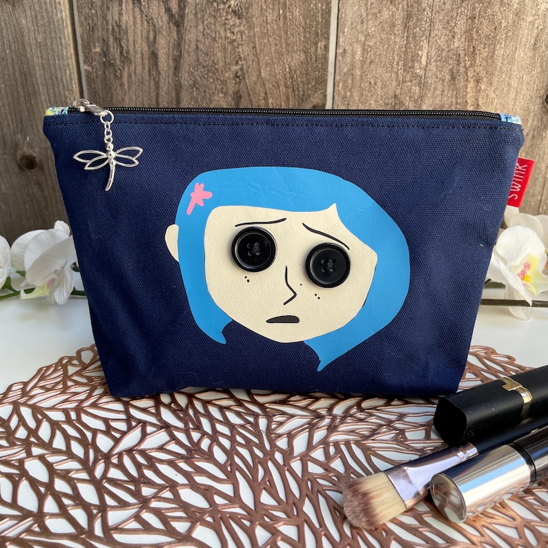 Puede incluir: Una bolsa de maquillaje de lona azul oscuro con una imagen impresa de una ni&ntilde;a con cabello azul y grandes botones negros para los ojos. La bolsa tiene una cremallera y un dije de lib&eacute;lula plateado.