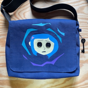 Puede incluir: Una bolsa de mensajero de lona azul oscuro con una ilustraci&oacute;n colorida de una ni&ntilde;a con cabello azul y ojos grandes. La ni&ntilde;a est&aacute; rodeada de un patr&oacute;n de remolino de azul, morado y rosa.