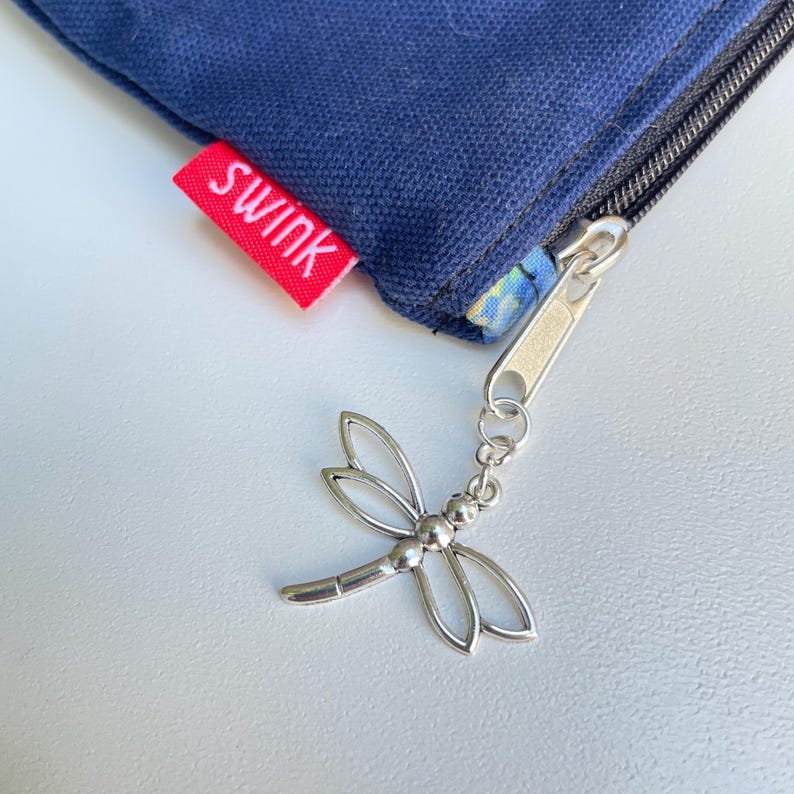 Puede incluir: Una peque&ntilde;a bolsa azul con cremallera con una etiqueta roja que dice "Swink" y un tirador de cremallera de lib&eacute;lula plateado.