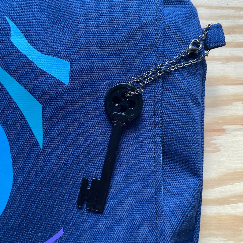 Puede incluir: Un llavero de llave negro con un dise&ntilde;o circular cuelga de una cadena plateada en una bolsa de lona azul oscuro.