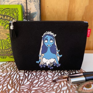 May include: Black canvas makeup bag with a zipper closure and a tassel pull. The bag features a colorful illustration of a blue-skinned bride with a white dress and a crown. The bride is surrounded by butterflies.