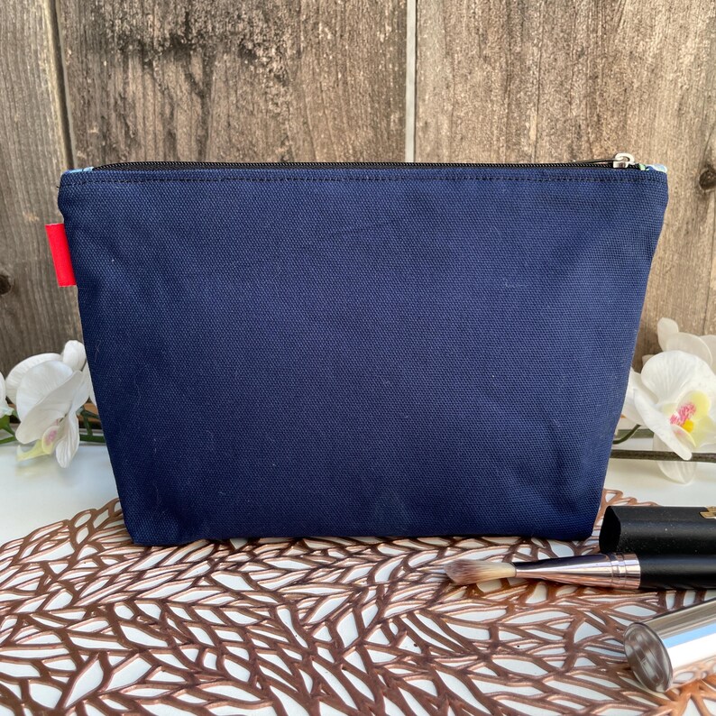 Puede incluir: Una bolsa de lona azul oscuro con cremallera y un tirador de cremallera rojo. La bolsa est&aacute; sobre una superficie decorativa.