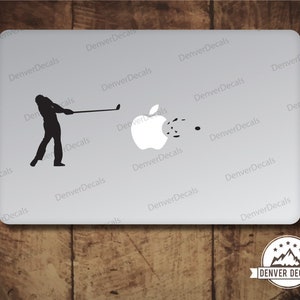 May include: A black silhouette of a golfer swinging a golf club, hitting an apple with a golf ball. The apple is white with a black stem and leaf. The background is a white laptop with the text "Denver Decals" repeated multiple times.