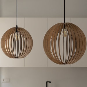 Pu&ograve; includere: Due lampade a sospensione sferiche realizzate con sottili listelli di legno curvati disposti a raggiera. Ogni lampada pende da un cavo nero e contiene una singola lampadina a vista, che diffonde una luce calda attraverso la struttura.