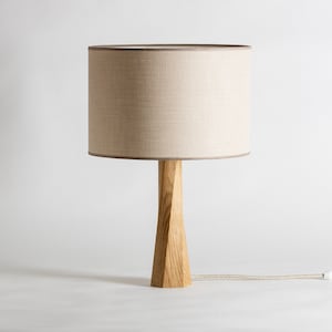 May include: A table lamp featuring a cylindrical beige fabric lampshade with a subtle texture and a light brown wooden base with a twisted, faceted geometric design. A fabric-covered power cord extends from the base to the right side.