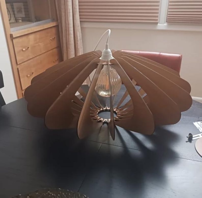 Puede incluir: Una l&aacute;mpara colgante que presenta una serie de listones de madera de color marr&oacute;n claro curvados, dispuestos en un patr&oacute;n circular y radial. La estructura rodea una bombilla de cristal transparente y estriado, suspendida de un cable blanco.