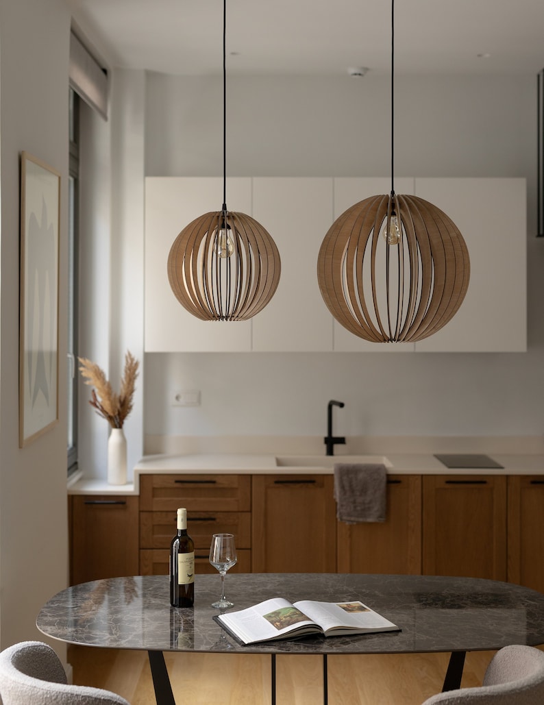 Pu&ograve; includere: Due lampade a sospensione sferiche realizzate con sottili listelli di legno curvati disposti a raggiera. Ogni lampada pende da un cavo nero e contiene una singola lampadina a vista, che diffonde una luce calda attraverso la struttura.