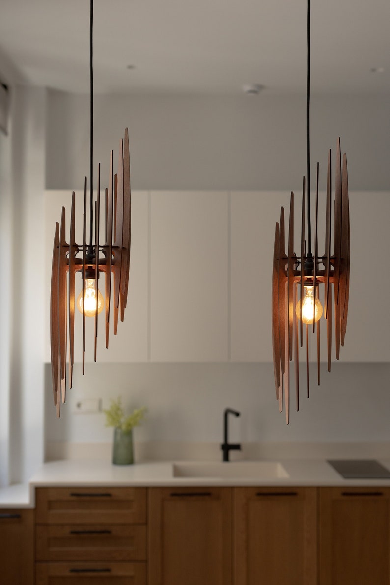 May include: Two modern wooden pendant lights with a geometric design hang from the ceiling above a kitchen counter. The lights are made of thin, flat pieces of wood arranged in a spiral pattern. Each light has a single light bulb.