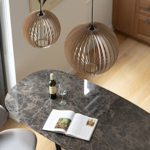 Pu&ograve; includere: Due lampade a sospensione sferiche realizzate con sottili listelli di legno chiaro disposti in un motivo verticale a gabbia. Ogni lampada pende da un sottile cavo nero e presenta una lampadina centrale visibile attraverso la struttura.