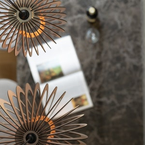 Pu&ograve; includere: Due lampade a sospensione sferiche realizzate con sottili listelli di legno chiaro disposti a raggiera. Ogni apparecchio presenta un supporto centrale circolare nero e un cavo scuro, creando un design a strati con struttura aperta.