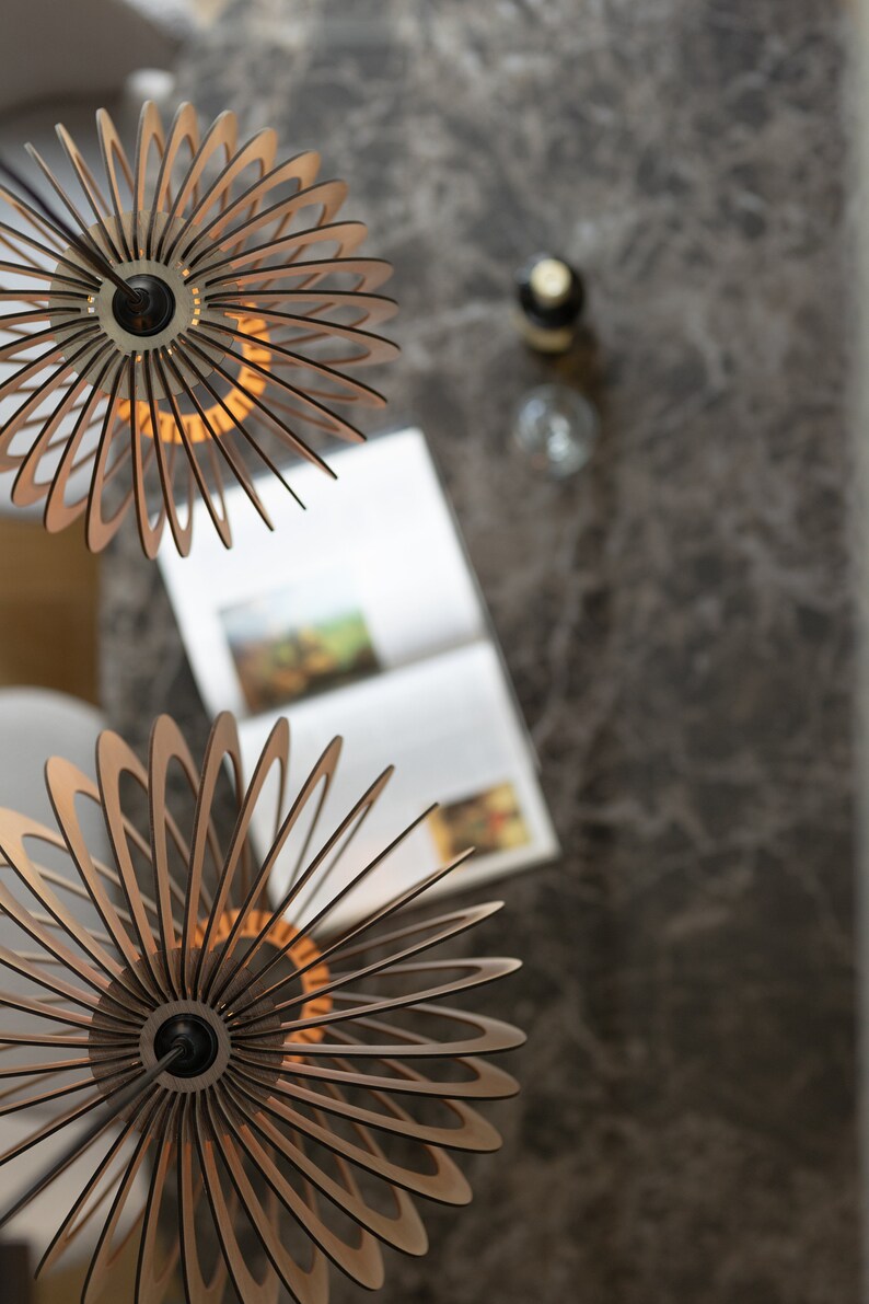 Pu&ograve; includere: Due lampade a sospensione sferiche realizzate con sottili listelli di legno chiaro disposti a raggiera. Ogni apparecchio presenta un supporto centrale circolare nero e un cavo scuro, creando un design a strati con struttura aperta.