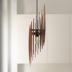 Op de afbeelding: Een hanglamp bestaande uit een reeks verticale, donkerbruine houten latten die in een gebogen, gelaagde formatie rond een centrale gloeilamp zijn gerangschikt. De lamp hangt aan een dun zwart snoer vanaf het plafond.