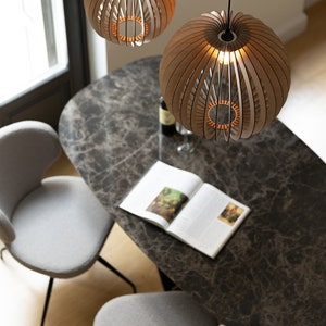 Pu&ograve; includere: Due lampade a sospensione sferiche realizzate con sottili listelli di legno curvati disposti a raggiera. Ogni lampada presenta una lampadina centrale che emette una luce calda, sospesa da un cavo nero sopra un tavolo da pranzo in marmo.