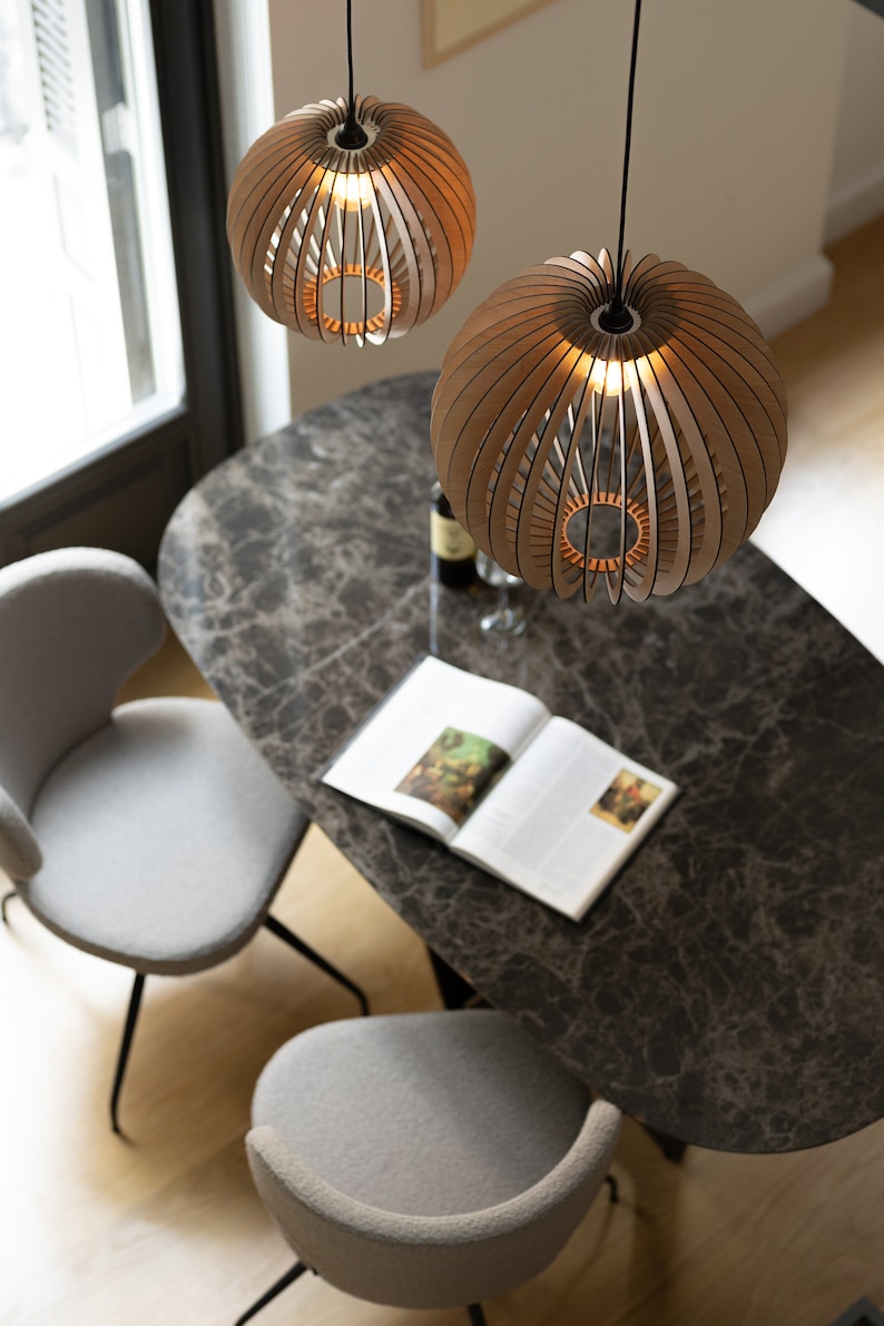 Pu&ograve; includere: Due lampade a sospensione sferiche realizzate con sottili listelli di legno curvati disposti a raggiera. Ogni lampada presenta una lampadina centrale che emette una luce calda, sospesa da un cavo nero sopra un tavolo da pranzo in marmo.
