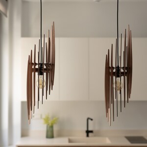 May include: Two modern wooden pendant lights with a geometric design hang from the ceiling above a kitchen counter. The lights have a warm, natural wood finish and are accented with black metal hardware.