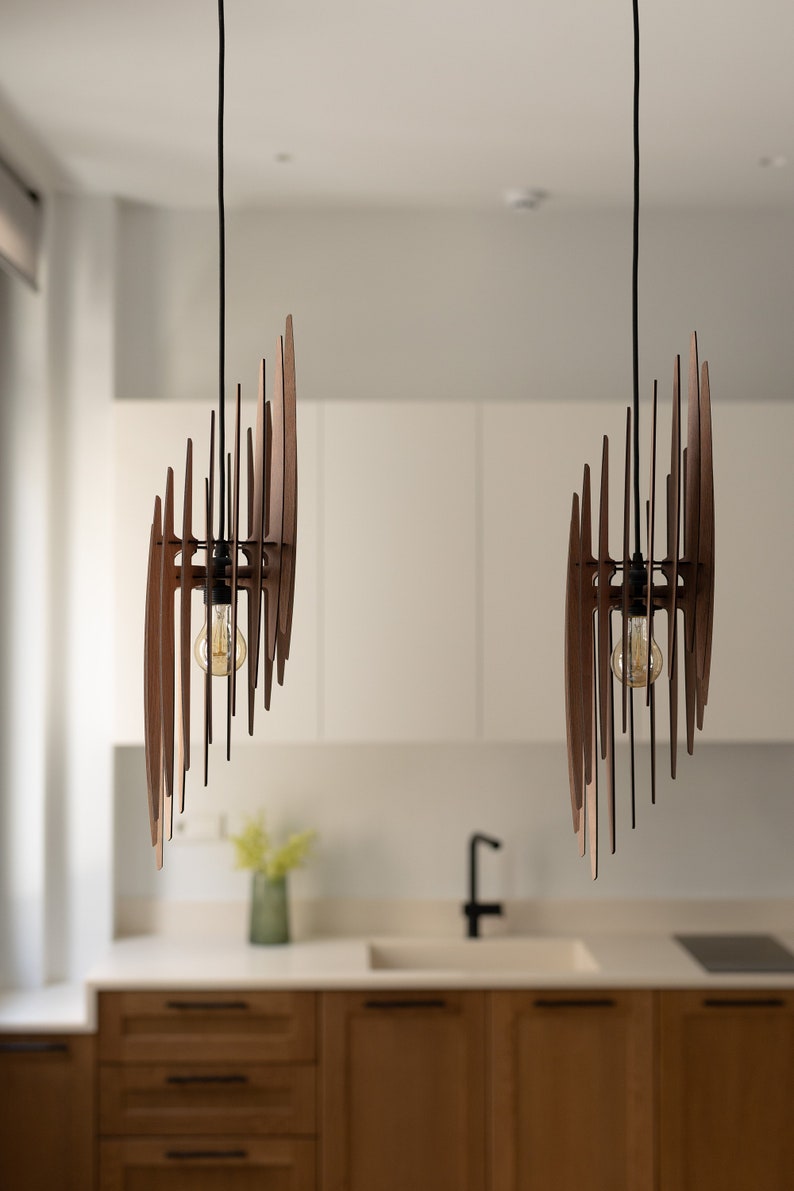 May include: Two modern wooden pendant lights with a geometric design hang from the ceiling above a kitchen counter. The lights have a warm, natural wood finish and are accented with black metal hardware.