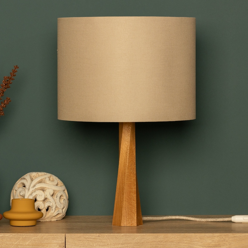 May include: A beige lampshade with a wooden base sits on a light brown surface. The lampshade is cylindrical and the base is a tall, slender, geometric shape.