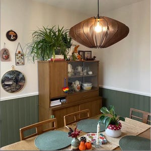 Puede incluir: Una l&aacute;mpara colgante de madera de color marr&oacute;n oscuro con un dise&ntilde;o redondeado de listones cuelga sobre una mesa de comedor. La l&aacute;mpara cuenta con una bombilla central que emite un brillo c&aacute;lido, proyectando luz a trav&eacute;s de los paneles.