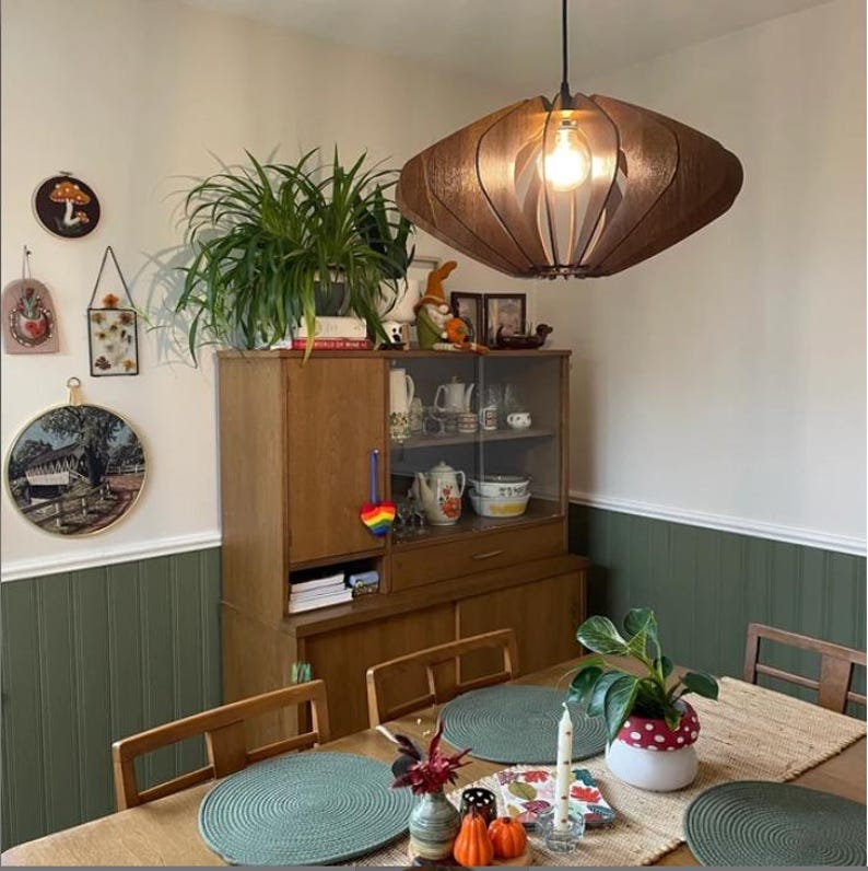 Puede incluir: Una l&aacute;mpara colgante de madera de color marr&oacute;n oscuro con un dise&ntilde;o redondeado de listones cuelga sobre una mesa de comedor. La l&aacute;mpara cuenta con una bombilla central que emite un brillo c&aacute;lido, proyectando luz a trav&eacute;s de los paneles.