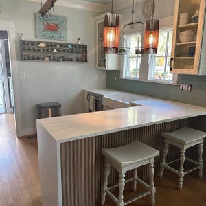 Puede incluir: Una cocina con encimera de cuarzo blanco sobre una base de listones de madera verticales, dos taburetes de madera clara y dos l&aacute;mparas colgantes cil&iacute;ndricas. Un estante de pared muestra frascos peque&ntilde;os y un letrero dice Seafood Market.