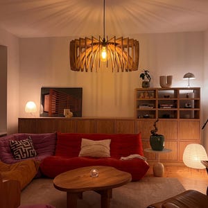 May include: A wooden pendant light fitting with a circular, slatted design hangs from the ceiling. The light features a warm-toned bulb that casts a soft glow, illuminating a living room with a red sofa, a wooden coffee table, and shelving.