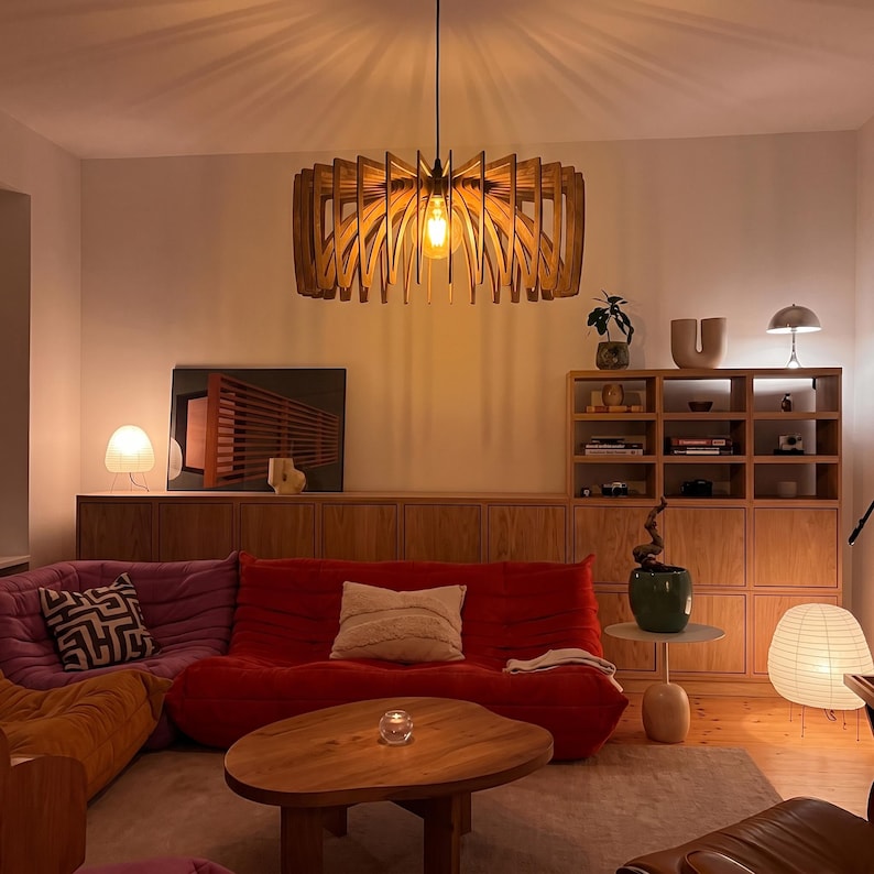 May include: A wooden pendant light fitting with a circular, slatted design hangs from the ceiling. The light features a warm-toned bulb that casts a soft glow, illuminating a living room with a red sofa, a wooden coffee table, and shelving.