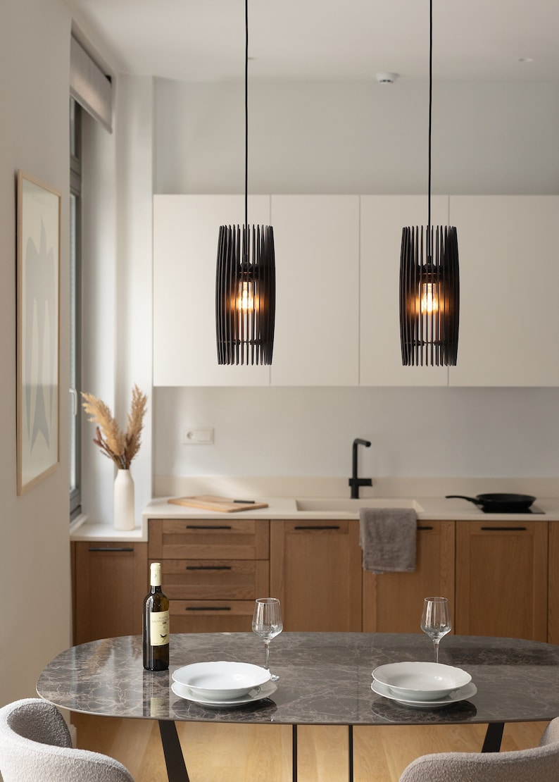 May include: Two cylindrical pendant lights featuring vertical black slats suspended from thin black cords. Each fixture houses a single glowing light bulb, casting a warm illumination downward over a dining area with a dark marble table.