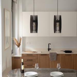 May include: Two cylindrical pendant lights with black vertical slats hanging over an oval marble dining table. The table is set with two white bowls, two clear wine glasses, and a wine bottle. The kitchen features light wood cabinets and white walls.