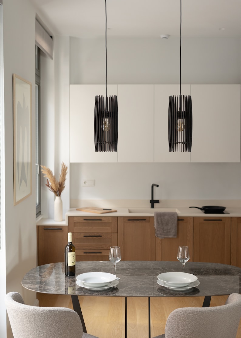 May include: Two cylindrical pendant lights with black vertical slats hanging over an oval marble dining table. The table is set with two white bowls, two clear wine glasses, and a wine bottle. The kitchen features light wood cabinets and white walls.