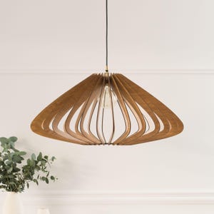 Può includere: Una lampada a sospensione con un ampio paralume a forma di piattino, costruito con listelli di legno marrone chiaro curvati e disposti a raggiera. Una singola lampadina in vetro trasparente è visibile al centro, sospesa da un sottile cavo nero.