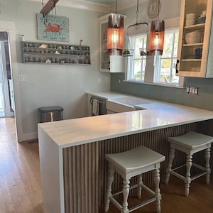 May include: A kitchen featuring a white quartz countertop with a vertical wood slat base, two light-colored wooden stools, and two cylindrical pendant lights. A wall-mounted shelf displays small jars, and a sign reads Seafood Market.