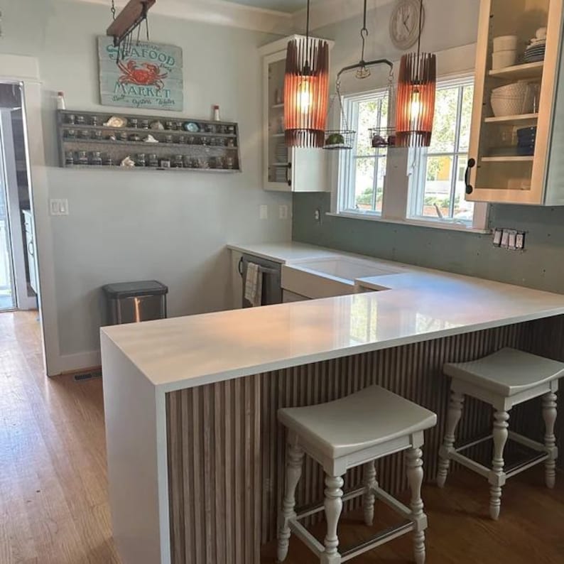 May include: A kitchen featuring a white quartz countertop with a vertical wood slat base, two light-colored wooden stools, and two cylindrical pendant lights. A wall-mounted shelf displays small jars, and a sign reads Seafood Market.