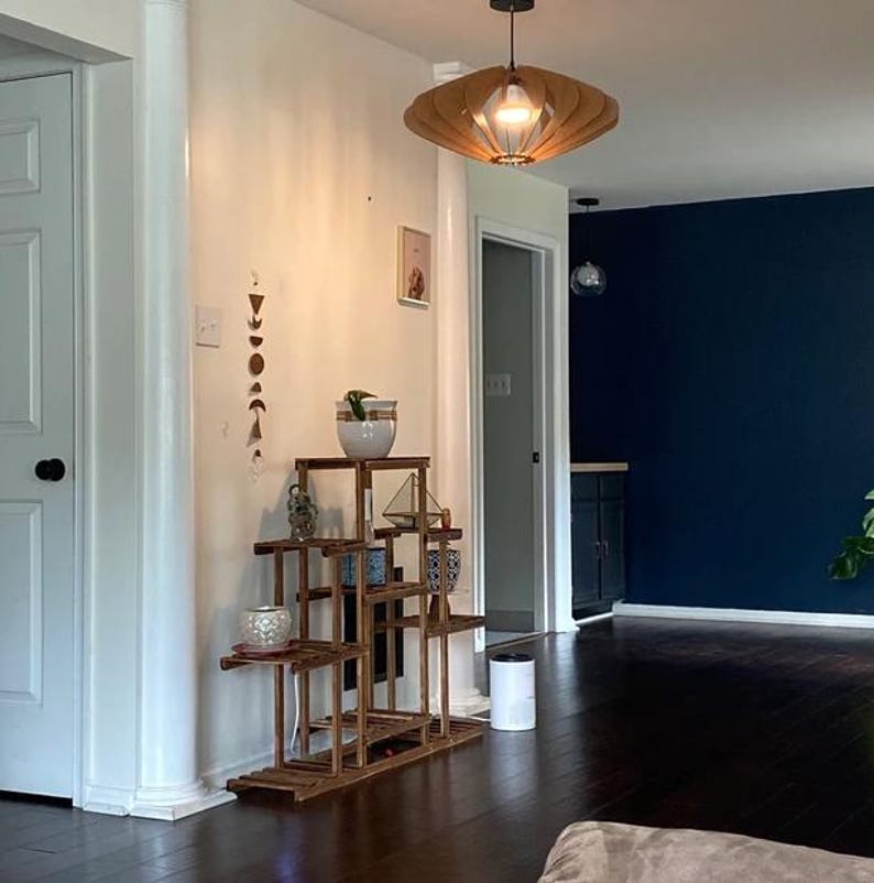 Puede incluir: Un soporte de madera para plantas de varios niveles con acabado marr&oacute;n oscuro, que cuenta con varios estantes a diferentes alturas. El soporte sostiene una maceta de cer&aacute;mica blanca con una peque&ntilde;a planta verde y un terrario geom&eacute;trico.