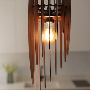 May include: A modern wooden pendant light fitting with a single light bulb. The light fitting is made of thin, vertical wooden slats that are arranged in a geometric pattern. The light bulb is a clear, Edison-style bulb that is turned on and glowing.