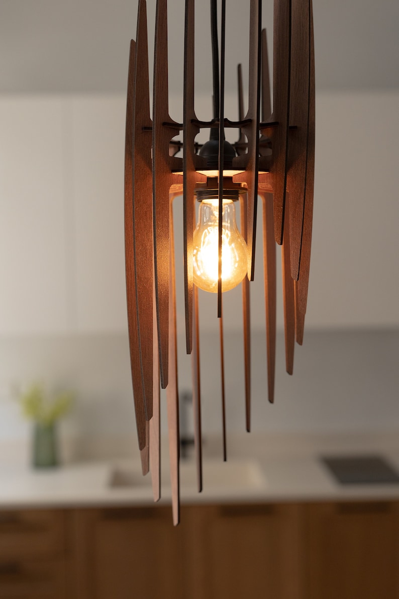 May include: A modern wooden pendant light fitting with a single light bulb. The light fitting is made of thin, vertical wooden slats that are arranged in a geometric pattern. The light bulb is a clear, Edison-style bulb that is turned on and glowing.