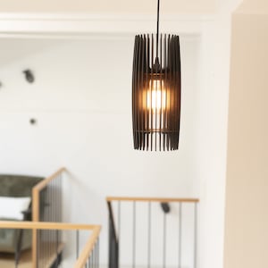 May include: A cylindrical pendant light fixture constructed from thin, vertical black slats arranged in a cage-like pattern. The fixture hangs from a black cord and contains a single glowing light bulb visible through the open slat design.
