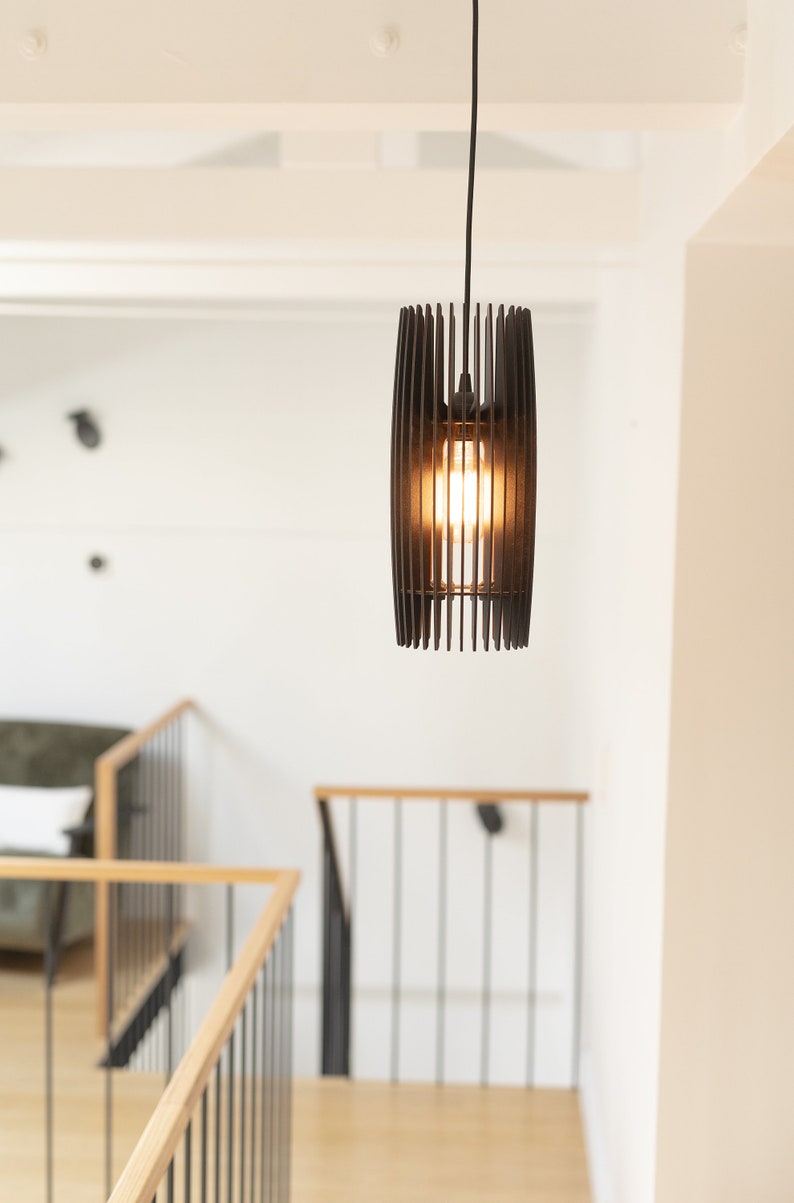May include: A cylindrical pendant light fixture constructed from thin, vertical black slats arranged in a cage-like pattern. The fixture hangs from a black cord and contains a single glowing light bulb visible through the open slat design.