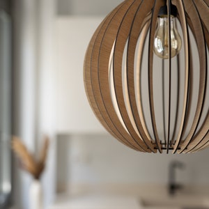 Pu&ograve; includere: Una lampada a sospensione sferica costruita con sottili listelli di legno marrone chiaro curvati e disposti in un motivo verticale. La struttura presenta un portalampada nero centrale con una lampadina in vetro trasparente.