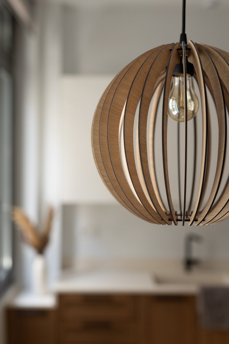 Pu&ograve; includere: Una lampada a sospensione sferica costruita con sottili listelli di legno marrone chiaro curvati e disposti in un motivo verticale. La struttura presenta un portalampada nero centrale con una lampadina in vetro trasparente.