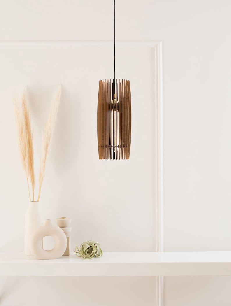 Puede incluir: Una l&aacute;mpara colgante cil&iacute;ndrica construida con listones de madera verticales de color marr&oacute;n oscuro. La luz cuelga de un cable negro fino y cuenta con una bombilla tubular central. El dise&ntilde;o crea una apariencia de marco abierto.