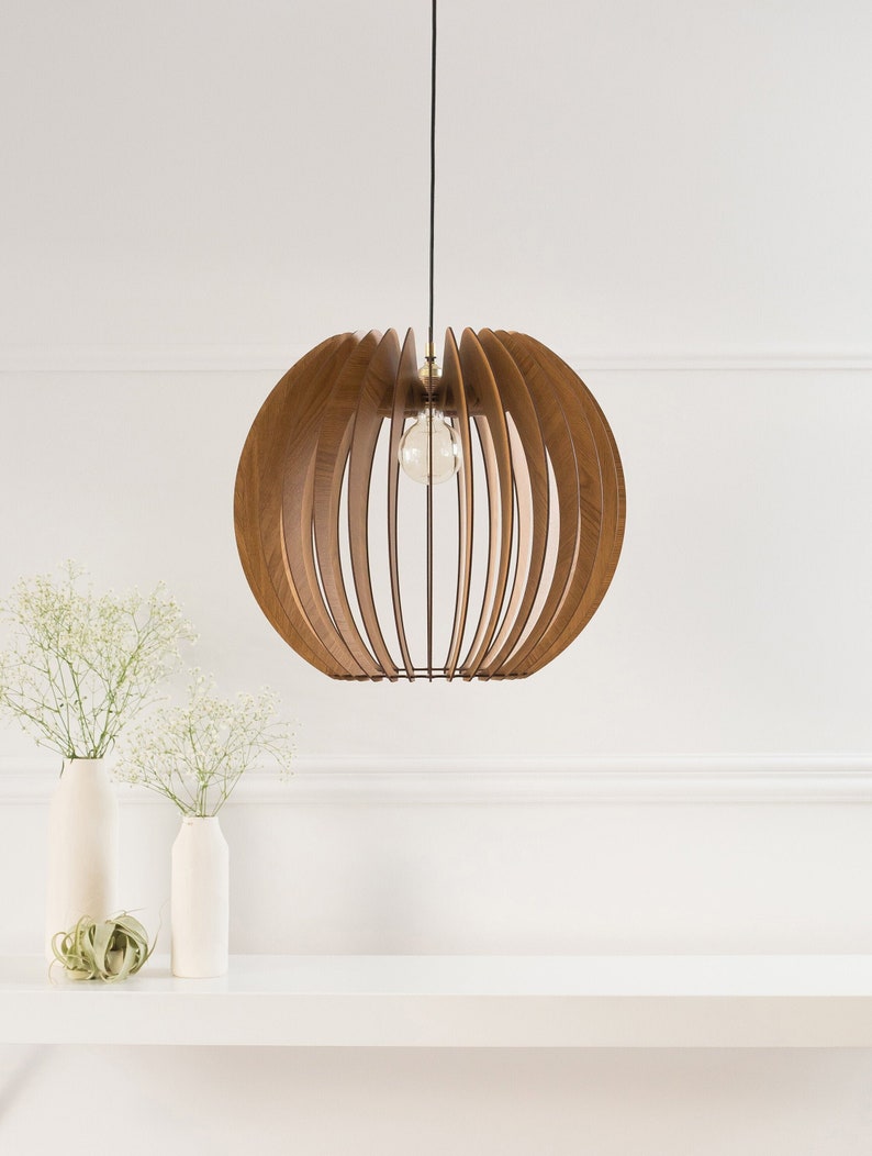 May include: A wooden pendant light fixture with a round, slatted shade. The shade is made of light brown wood and has a natural finish. The light fixture is hanging from a black cord and is illuminated by a single light bulb.