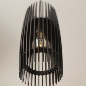 May include: A dark brown pendant light fixture featuring a cylindrical cage design composed of vertical slats. The structure is suspended from a thin cord and contains a single light bulb visible through the open gaps between the wooden slats.