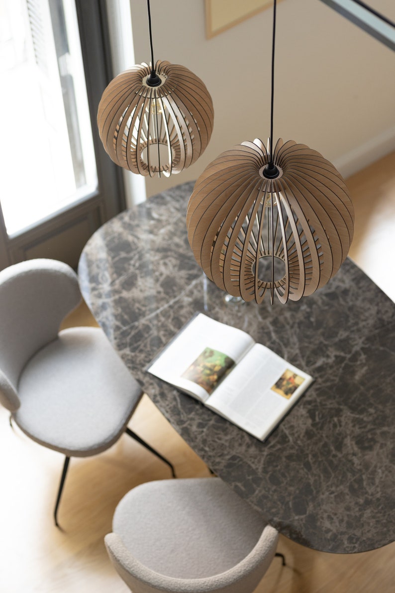 Pu&ograve; includere: Due lampade a sospensione sferiche realizzate con sottili listelli di legno marrone chiaro disposti a raggiera. Ogni lampada presenta un'apertura centrale ed &egrave; sospesa tramite un sottile cavo nero fissato al soffitto sopra un tavolo.