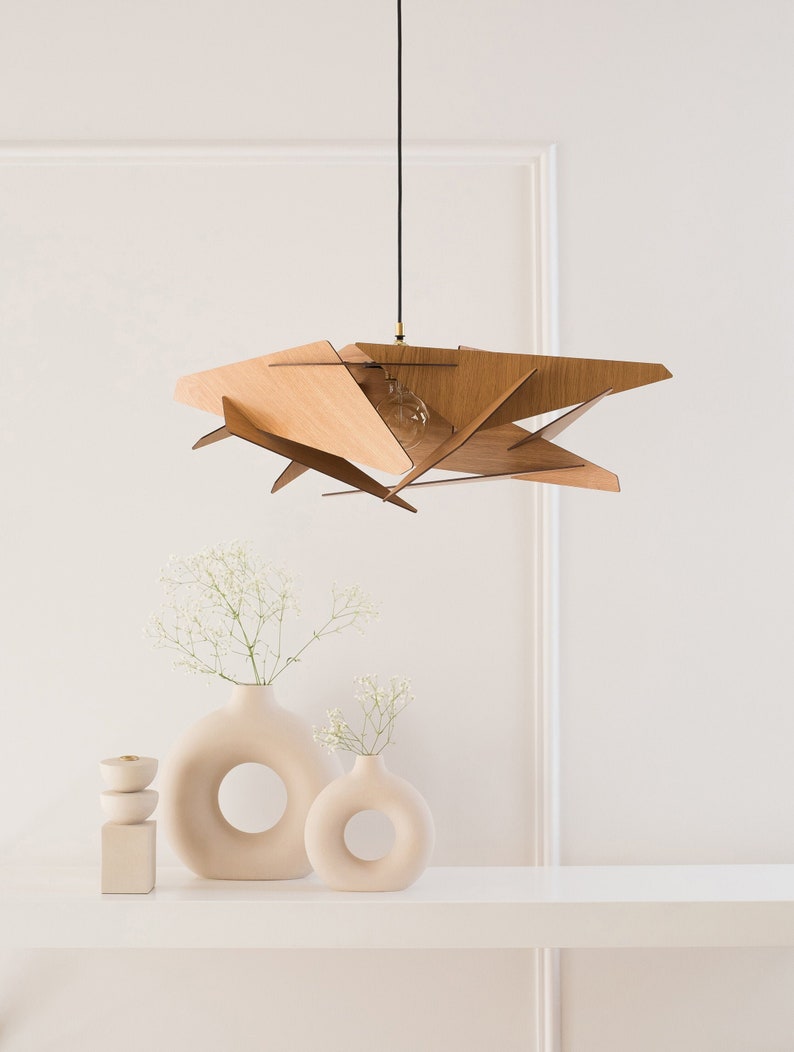 May include: A wooden pendant light fixture with a geometric design hangs from a black cord. Two ceramic vases with circular cutouts, one larger than the other, are on a white shelf. Both vases are filled with white flowers.
