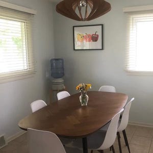 Puede incluir: Un comedor con una mesa ovalada de madera marr&oacute;n oscuro con patr&oacute;n de espiga, rodeada de seis sillas blancas. Un florero de vidrio con flores amarillas descansa sobre la mesa. Una l&aacute;mpara colgante de madera cuelga junto a un cuadro.