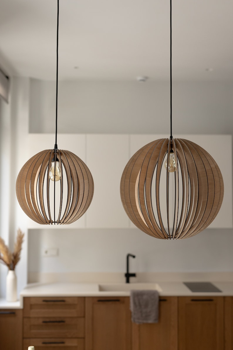 Pu&ograve; includere: Due lampade a sospensione sferiche realizzate con sottili listelli di legno marrone chiaro curvati e disposti a raggiera. Ogni lampada pende da un cavo nero e contiene una singola lampadina a vista al centro della struttura in legno.