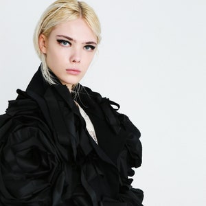 Puede incluir: Una mujer con un vestido negro con grandes mangas con volantes. El vestido est&aacute; hecho de una tela suave y fluida y tiene una silueta voluminosa y dram&aacute;tica.