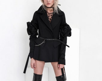 May include: A black blazer with a chain belt and large, ruffled sleeves. The blazer is worn over a black sheer top with polka dots. The model is wearing black knee-high boots.
