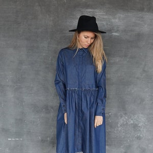 May include: A woman wearing a dark blue denim shirt dress with a gathered waist and long sleeves. She is wearing a black hat and black ankle boots.