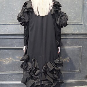 Puede incluir: Un vestido negro con falda larga y grandes volantes. El vestido tiene un corpi&ntilde;o ajustado y mangas largas. La modelo lleva botas negras.
